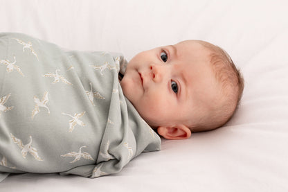 Baby lying wrapped in Swan Flight Swaddle Blanket by Burrow and Be