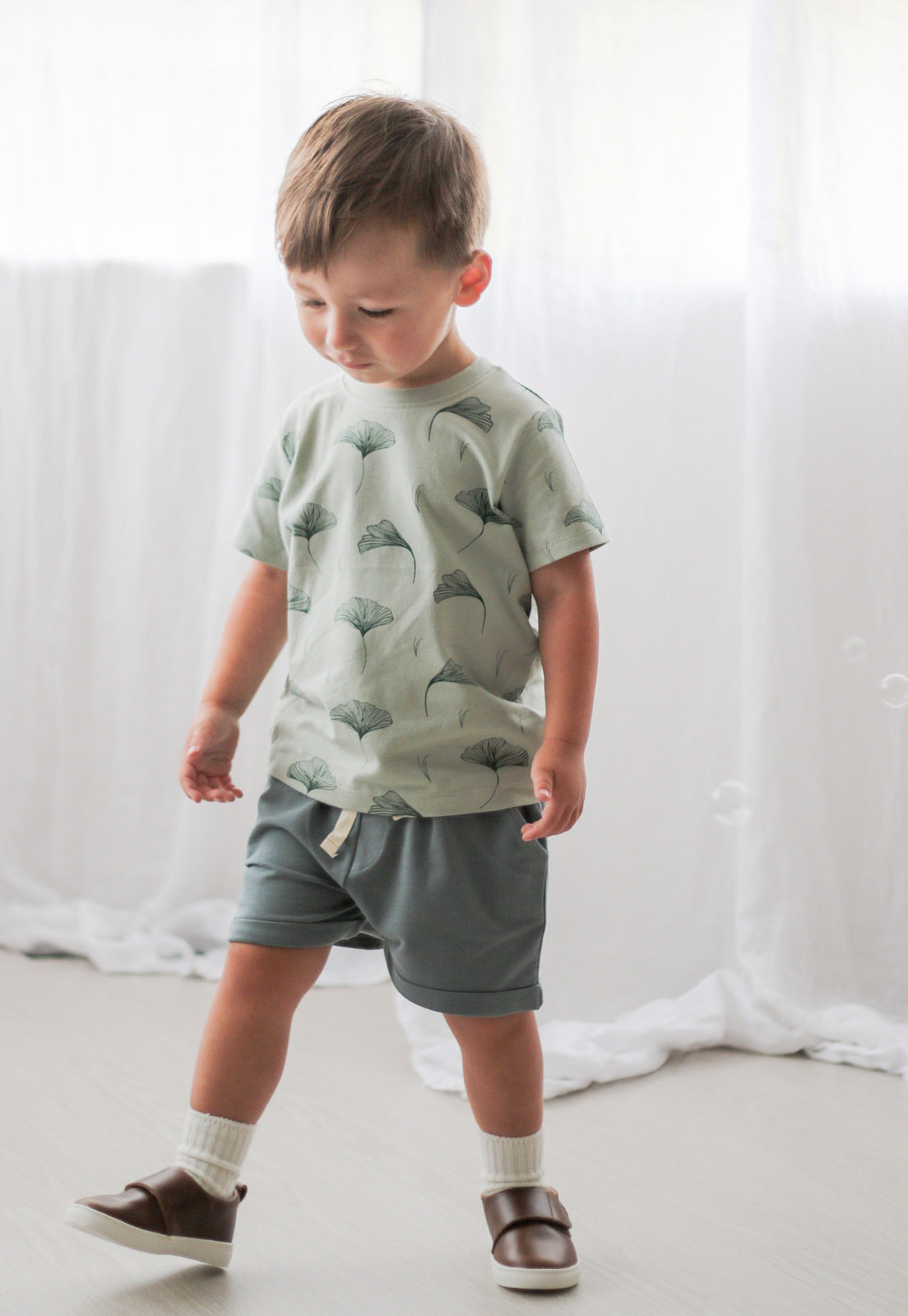 Boy posing in front of the camera wearing Shoes by Wanders, Ginkgo T-shirt and Terry Knit Baby Shorts in Steel against white curtains, all designed by Burrow and Be