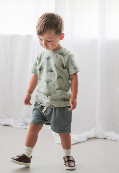 Boy posing in front of the camera wearing Shoes by Wanders, Ginkgo T-shirt and Terry Knit Baby Shorts in Steel against white curtains, all designed by Burrow and Be